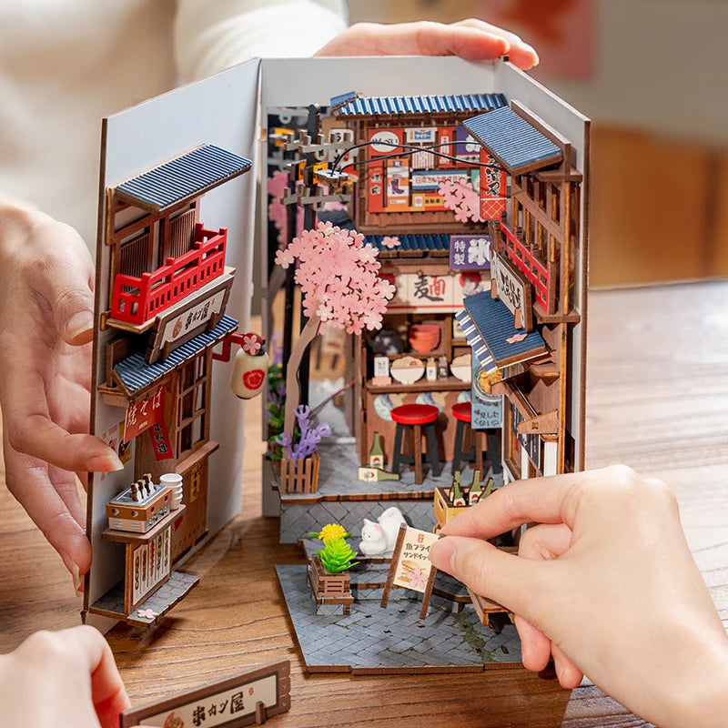 Miniature model of a Japanese street scene with buildings and cherry blossoms, held open by hands.